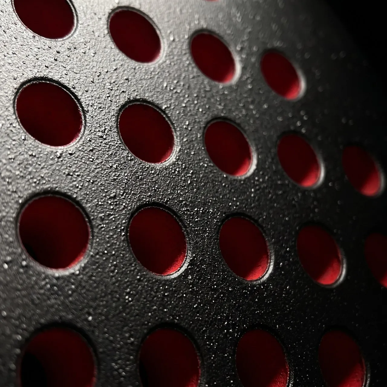 Macro du tamis d'une raquette SCUD Black Impact B3 : les perforations aux parois rouges et, entre elles, le grain du sablage fait-main sur la surface noire